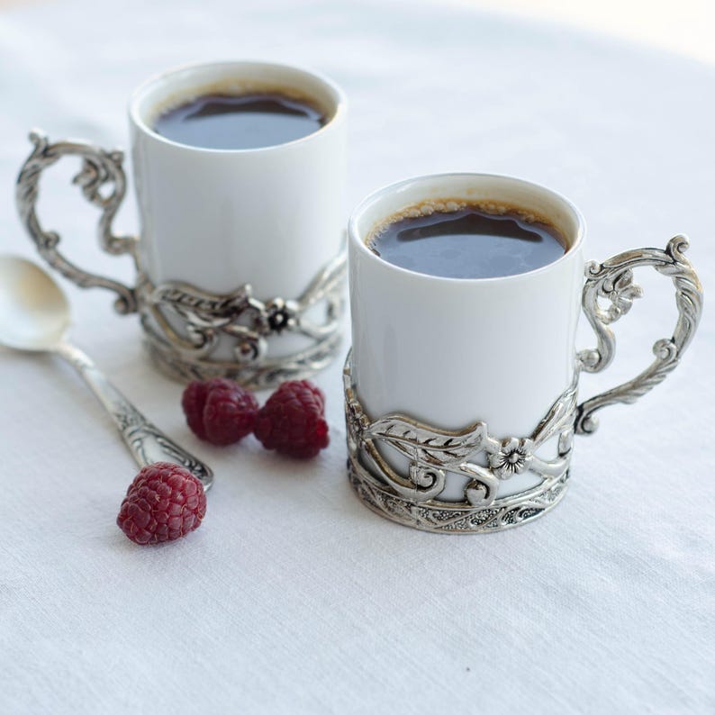 May include: Two white ceramic coffee mugs filled with dark liquid, each held by ornate silver-coloured holders. A silver spoon and several red raspberries are next to the mugs on a white surface. The mugs have decorative handles.