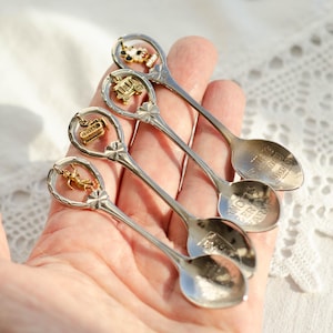 Puede incluir: Cuatro cucharas de recuerdo plateadas con adornos decorativos. Cada cuchara tiene un adorno diferente: una vaca, un tren, una diligencia y una figura dorada. Las cucharas tienen texto grabado. Las cucharas se sostienen en una mano.