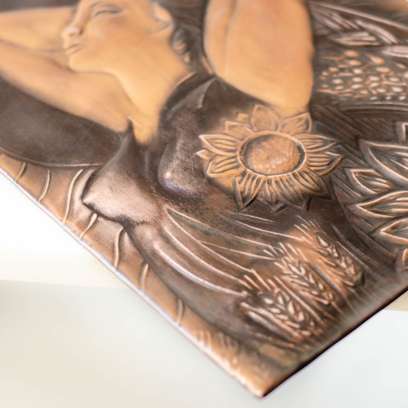 May include: A copper-toned wall plaque with a raised relief design of a woman with a sunflower and wheat stalks. The woman's face is serene and her eyes are closed.