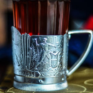 May include: A silver metal cup with an intricate design featuring a figure holding a torch. The cup is filled with a dark red liquid and has a handle.
