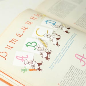 May include: A vintage children's book with a colorful alphabet illustration. The letters A, B, and C are shown with cartoon characters holding various foods, including carrots, wheat, and grapes. The text on the page reads "Витамины способствуют росту и правильному развитию детей, повышают их выносливость и сопротивляемость организма различным заболеваниям."