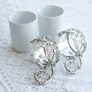 May include: Two white ceramic cups and two ornate silver-coloured cup holders with floral and leaf designs. The cup holders have a decorative handle and are placed on a white lace tablecloth. The cups are cylindrical.
