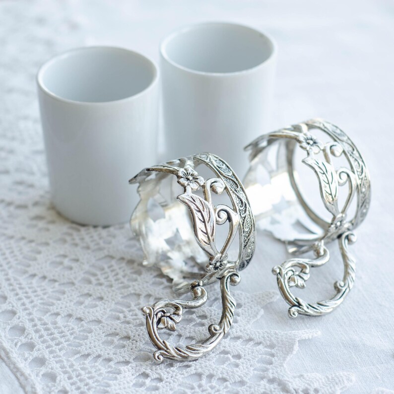 May include: Two white ceramic cups and two ornate silver-coloured cup holders with floral and leaf designs. The cup holders have a decorative handle and are placed on a white lace tablecloth. The cups are cylindrical.