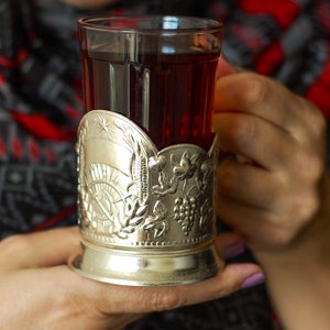 May include: A silver tea cozy with an ornate design, holding a glass of red tea. The cozy features a star, wheat, and grapes.