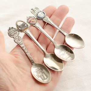 May include: Four silver souvenir spoons with detailed designs. Each spoon features a different emblem, including a shield, an eagle, and a building. The spoons are held in a hand, showcasing their intricate craftsmanship and collectible nature.
