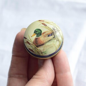 Puede incluir: Una caja de baratijas redonda y decorativa con una ilustraci&oacute;n detallada de un pato real en un entorno pantanoso. La caja tiene un fondo color crema, un borde azul y una base de metal dorado. El pato tiene plumas verdes, marrones y azules.