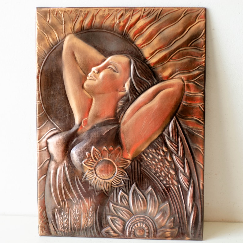 May include: A copper wall sculpture of a woman with her arms raised, surrounded by sunflowers and wheat. The sculpture has a textured, hammered finish.