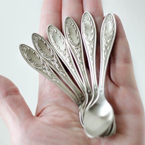 Puede incluir: Un juego de seis cucharaditas de plata con diseños ornamentados, extendidas en una mano.