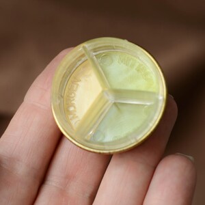 Puede incluir: Un pastillero redondo y segmentado con un borde dorado y un interior transparente dividido. Los compartimentos est&aacute;n etiquetados con palabras alemanas. El pastillero se sostiene en una mano.