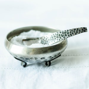 May include: A silver salt cellar with a hammered finish and a matching spoon. The cellar has three small feet and is filled with white salt.