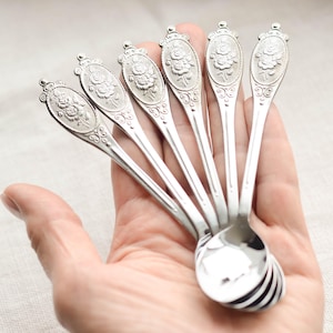 Könnte beinhalten: Ein Set aus sechs silberfarbenen Löffeln. Jeder Löffel hat ein ovales Design mit einer Rose in der Mitte und dekorativen Verzierungen. Die Löffel werden in einer Hand gehalten, was ihre polierte Oberfläche und die feinen Details zeigt.