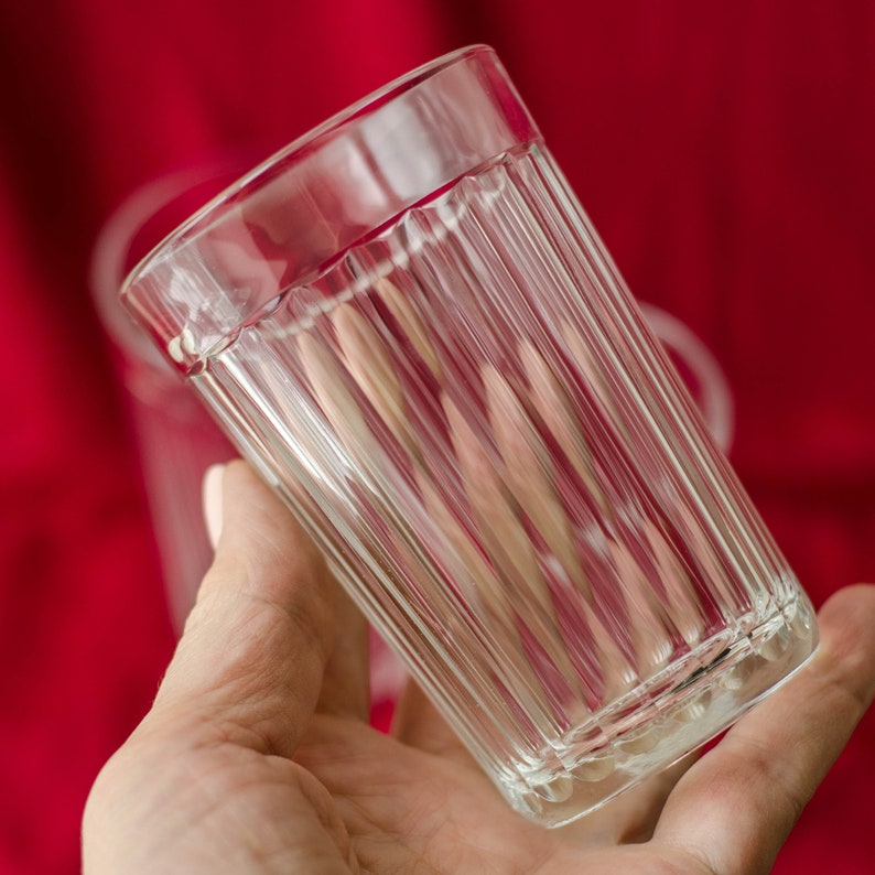 Op de afbeelding: Een helder drinkglas met een reeks verticale ribbels langs de buitenwanden. De cilindrische beker heeft een gladde, dikke rand en een platte bodem, met een transparant ontwerp dat het licht door het gestructureerde oppervlak laat schijnen.