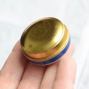 Puede incluir: Una peque&ntilde;a lata redonda vintage con tapa dorada, banda color cobre y base azul. La lata se sostiene en una mano. La lata parece ser de metal.