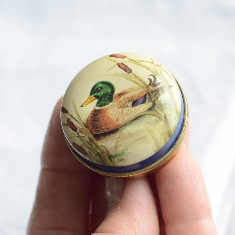 Puede incluir: Una caja de baratijas redonda y decorativa con una ilustraci&oacute;n detallada de un pato real en un entorno de marisma. El pato tiene plumaje verde, marr&oacute;n y azul, con un pico amarillo. La caja tiene un borde azul y una base dorada.