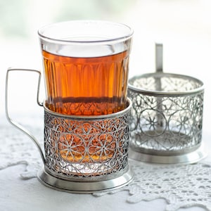 May include: A glass of amber-colored tea in a decorative silver filigree cup holder with a handle. Beside it is a matching, empty cup holder. The cup and holders sit on a white, lacy surface.