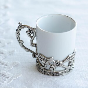 May include: A white ceramic mug with a decorative silver-coloured metal handle and base. The handle has an ornate, floral design. The base of the mug is encircled by a matching metal band with a similar floral pattern.