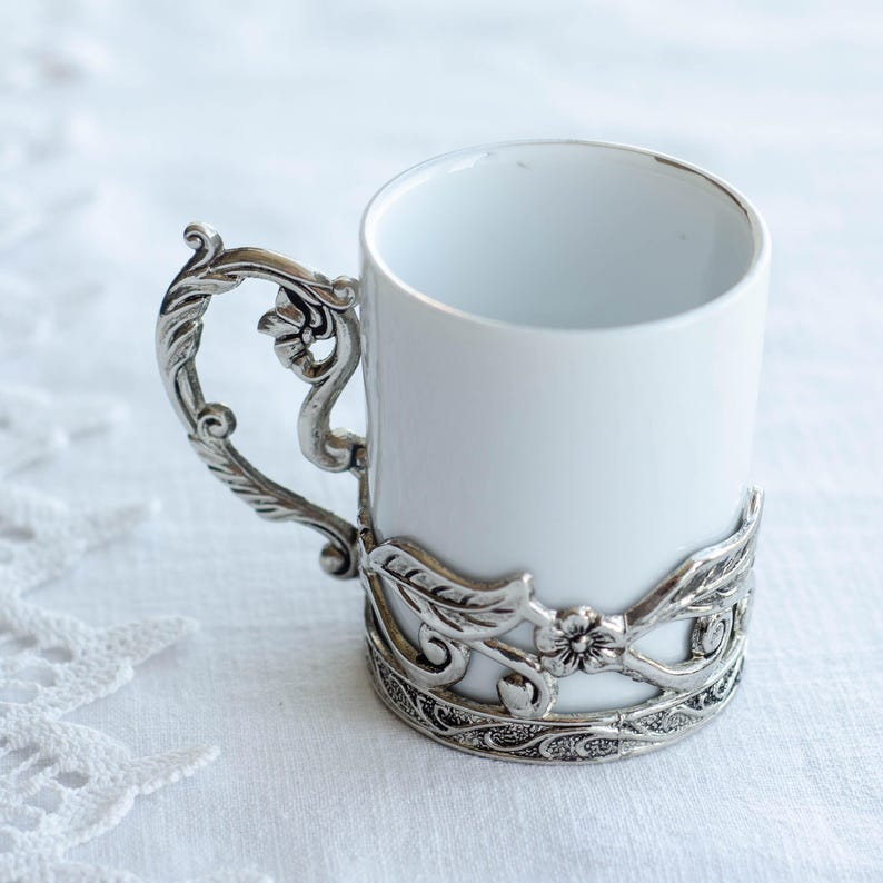 May include: A white ceramic mug with a decorative silver-coloured metal handle and base. The handle has an ornate, floral design. The base of the mug is encircled by a matching metal band with a similar floral pattern.