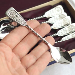 Puede incluir: Juego de cucharas plateadas con diseños florales en los mangos y tallos retorcidos. Las cucharas se presentan en una caja con un interior morado. Una cuchara se sostiene en una mano.