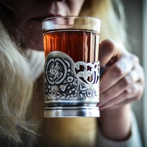 Op de afbeelding: Een theeglas van helder glas in een decoratieve zilverkleurige metalen houder. De metalen houder is voorzien van een reli&euml;f met een rond medaillon van een ruiter te paard, omringd door gedetailleerde bloem- en bladmotieven aan de onderzijde.