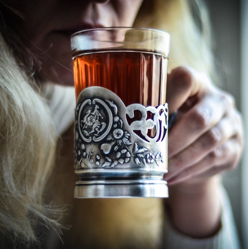 Op de afbeelding: Een theeglas van helder glas in een decoratieve zilverkleurige metalen houder. De metalen houder is voorzien van een reli&euml;f met een rond medaillon van een ruiter te paard, omringd door gedetailleerde bloem- en bladmotieven aan de onderzijde.