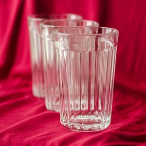 Op de afbeelding: Drie heldere glazen drinkbekers op een rij, elk met een verticaal geribbelde textuur langs de zijkanten en een gladde, ronde rand. De glazen zijn transparant en cilindervormig, met een consistent patroon van verticale ribbels.