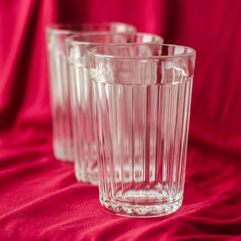 Op de afbeelding: Drie heldere glazen drinkbekers op een rij, elk met een verticaal geribbelde textuur langs de zijkanten en een gladde, ronde rand. De glazen zijn transparant en cilindervormig, met een consistent patroon van verticale ribbels.
