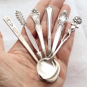 Puede incluir: Una colección de cucharas de plata con diseños ornamentados en los mangos, incluyendo patrones florales, de hojas y geométricos. Las cucharas están dispuestas en forma de abanico, mostrando sus intrincados detalles y superficies pulidas. Las cucharas se sostienen en una mano.