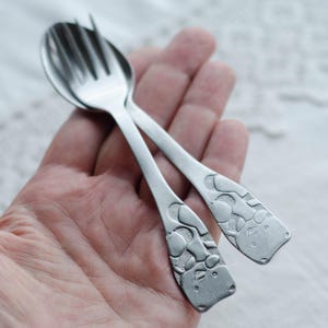 Peut inclure: Une paire de petits ustensiles argentés : une fourchette et une cuillère. Les manches présentent un motif texturé. Les ustensiles sont tenus dans une main, montrant leur taille et leurs détails. La fourchette a quatre dents.