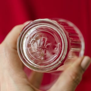 Op de afbeelding: Een helder drinkglas met een geribbelde buitenkant. De onderkant van het glas is voorzien van een reli&euml;flogo bestaande uit een gestileerde kroonvorm boven het getal 800 en het getal 23, die direct in de bodem van het glas zijn gegoten.