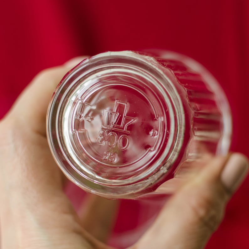Op de afbeelding: Een helder drinkglas met een geribbelde buitenkant. De onderkant van het glas is voorzien van een reli&euml;flogo bestaande uit een gestileerde kroonvorm boven het getal 800 en het getal 23, die direct in de bodem van het glas zijn gegoten.