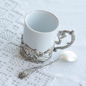 May include: A white ceramic cup with a silver filigree holder and handle, and a matching spoon. The cup and spoon feature an ornate, vintage design, set against a white lace background.
