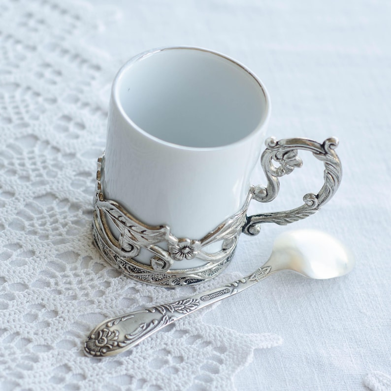 May include: A white ceramic cup with a silver filigree holder and handle, and a matching spoon. The cup and spoon feature an ornate, vintage design, set against a white lace background.