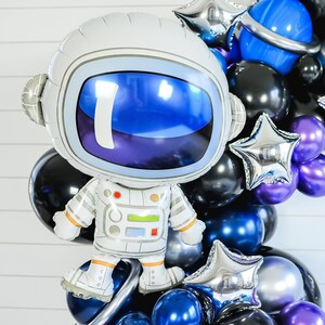 May include: A white astronaut balloon with a blue visor and a silver star balloon. The balloons are part of a balloon arch with other balloons in black, blue, silver, and purple.