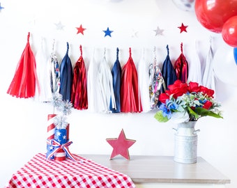 May include: Red, white, and blue tassel garland with stars hanging on a white wall. A red, white, and blue checkered tablecloth is on a white table with a red wooden star and a vase with red, white, and blue flowers.