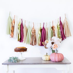 May include: A white table with a brown cake on a white cake stand, two pumpkins, and a bouquet of white flowers. A garland of tissue paper tassels in shades of gold, green, and burgundy hangs on the wall behind the table.