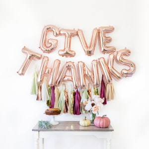 May include: Rose gold foil balloons spelling "GIVE THANKS" with a white background. The balloons are decorated with tassels in various shades of pink, cream, and green. A small table with a cake and pumpkins sits in front of the balloons.