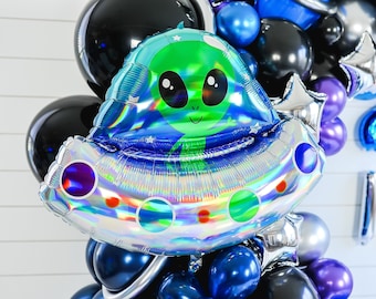 Könnte beinhalten: Ein silberner und blauer, schimmernder Ballon in Form eines Raumschiffs mit einem grünen Außerirdischen darin. Der Ballon ist Teil eines Ballonbogens mit anderen Ballons in Schwarz, Blau, Lila und Silber.