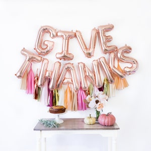 May include: Rose gold foil balloons spelling "GIVE THANKS" with pink, white, and gold tissue paper streamers hanging from the balloons. A small table with a cake, pumpkins, and flowers sits in front of the balloons.