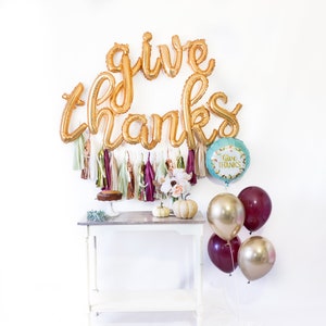 May include: Gold foil balloons spelling "give thanks" with a teal balloon with the text "Give Thanks" and burgundy and gold balloons. There are also gold and burgundy tassels and a small white table with a cake on it.