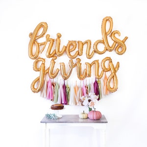 May include: Gold foil balloons spelling "Friendsgiving" with a garland of colorful tassels hanging below. A small table with a cake and two pumpkins sits in front of the balloons.
