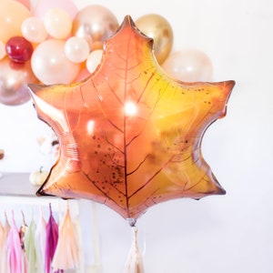 May include: A large, orange and yellow foil balloon shaped like a maple leaf. The balloon has a brown tassel attached to the bottom.