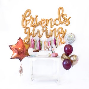 May include: Gold foil balloon letters spelling "Friendsgiving" with a white background. There are also several balloons in burgundy and gold, a leaf-shaped balloon, and a small white balloon with the text "Happy Thanksgiving".