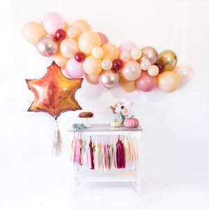 May include: A fall-themed party table setting with a balloon arch in shades of peach, pink, and gold, a large leaf-shaped balloon, a small cake, and two pumpkins. The table is decorated with a garland of pink, peach, and burgundy tassels.