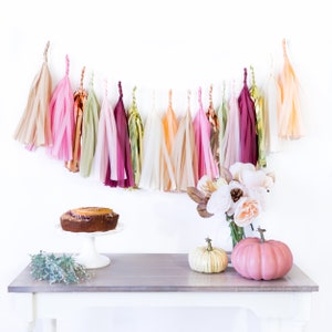 May include: A table setting with a cake, pumpkins, and a bouquet of white flowers. A garland of tissue paper tassels in various shades of pink, peach, and green hangs above the table.
