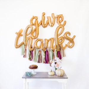 May include: Gold foil balloons spelling "give thanks" with a garland of burgundy, beige, and gold tassels hanging below. A white table with a cake and pumpkins on it.