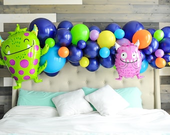 May include: A bed with a headboard decorated with a balloon arch featuring blue, green, yellow, orange, and purple balloons. Two monster-shaped balloons are on either side of the arch.