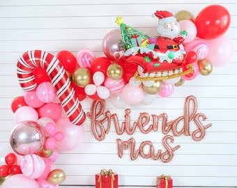 May include: A Christmas balloon arch with red, pink, and gold balloons. The arch includes a Santa Claus balloon riding in a sleigh, a candy cane balloon, and a balloon that says "Friendsmas".