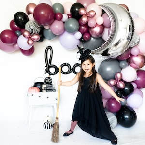 May include: A young girl dressed as a witch holds a broom in front of a black 'boo' sign and a backdrop of purple, pink, and silver balloons. She is wearing a black dress, a witch hat, and striped tights.