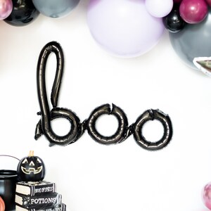 May include: Black balloon letters spelling "boo" with a black cauldron, a pumpkin, and a broom in the bottom left corner. The balloons are surrounded by a garland of purple, pink, and gray balloons.