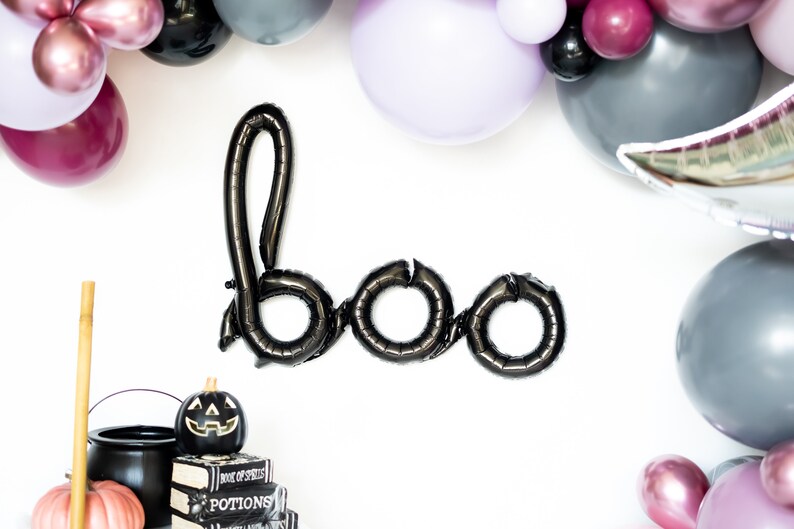 May include: Black balloon letters spelling "boo" with a black cauldron, a pumpkin, and a broom in the bottom left corner. The balloons are surrounded by a garland of purple, pink, and gray balloons.